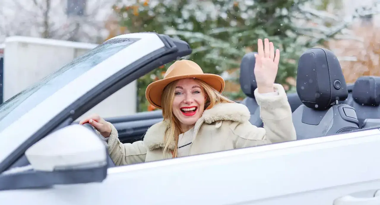 Fröhliche Frau winkt aus einem weißen Cabrio im Winter, während Schneeflocken fallen.