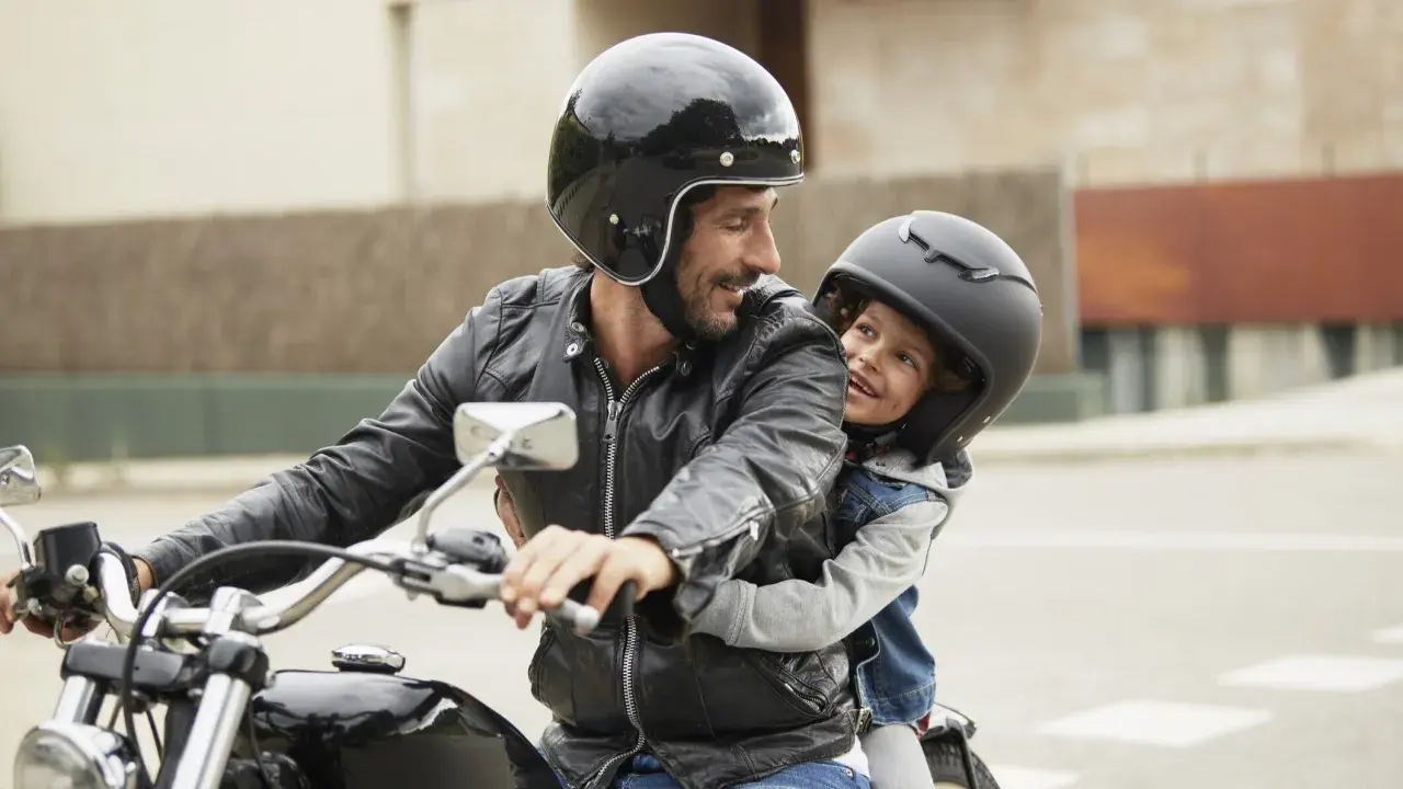 Ein Mann mit Bart und schwarzer Lederjacke nimmt ein Kind auf seinem Motorrad mit. Beide tragen Helme und lächeln sich an.