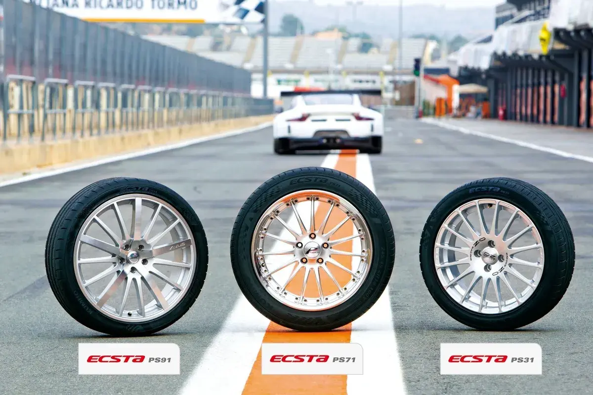 Drei Kumho Ecsta PS71 Reifen stehen auf einer Rennstrecke, bereit für einen Test. Ein weißer Sportwagen fährt im Hintergrund.