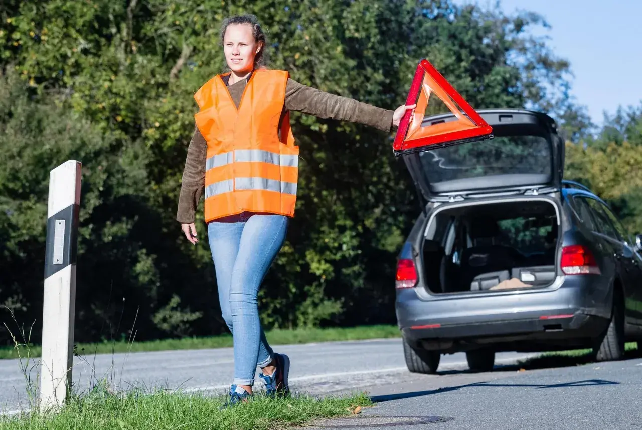 Frau mit Warnweste und Warndreieck neben offenem Kofferraum. Sie bereitet sich auf das auto aufbocken vor, um das Fahrzeug zu reparieren.
