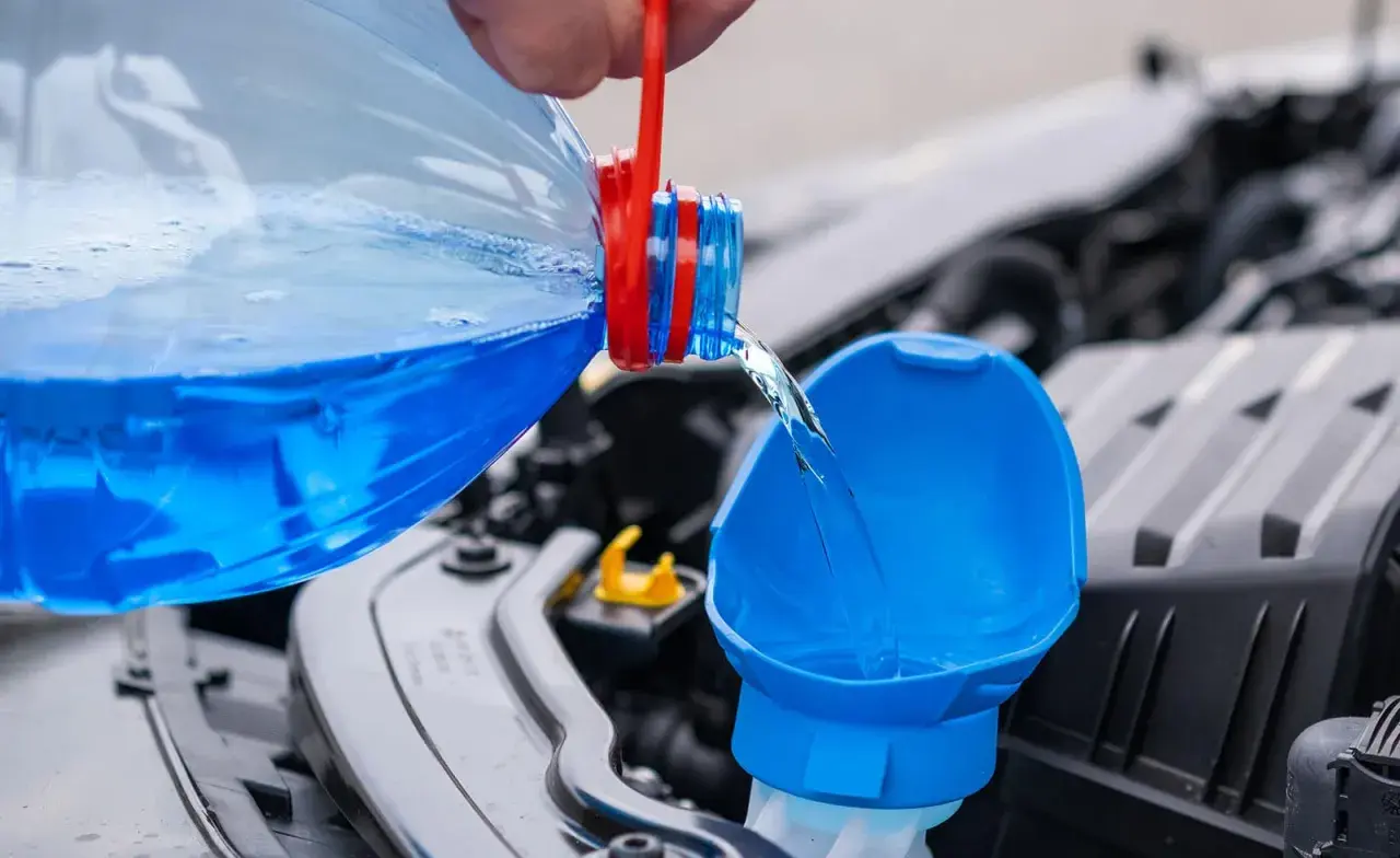 Blauer Scheibenreiniger wird aus einer Flasche in den Einfüllstutzen gegossen. Perfekt für das Scheibenwischwasser Sommer selber machen.