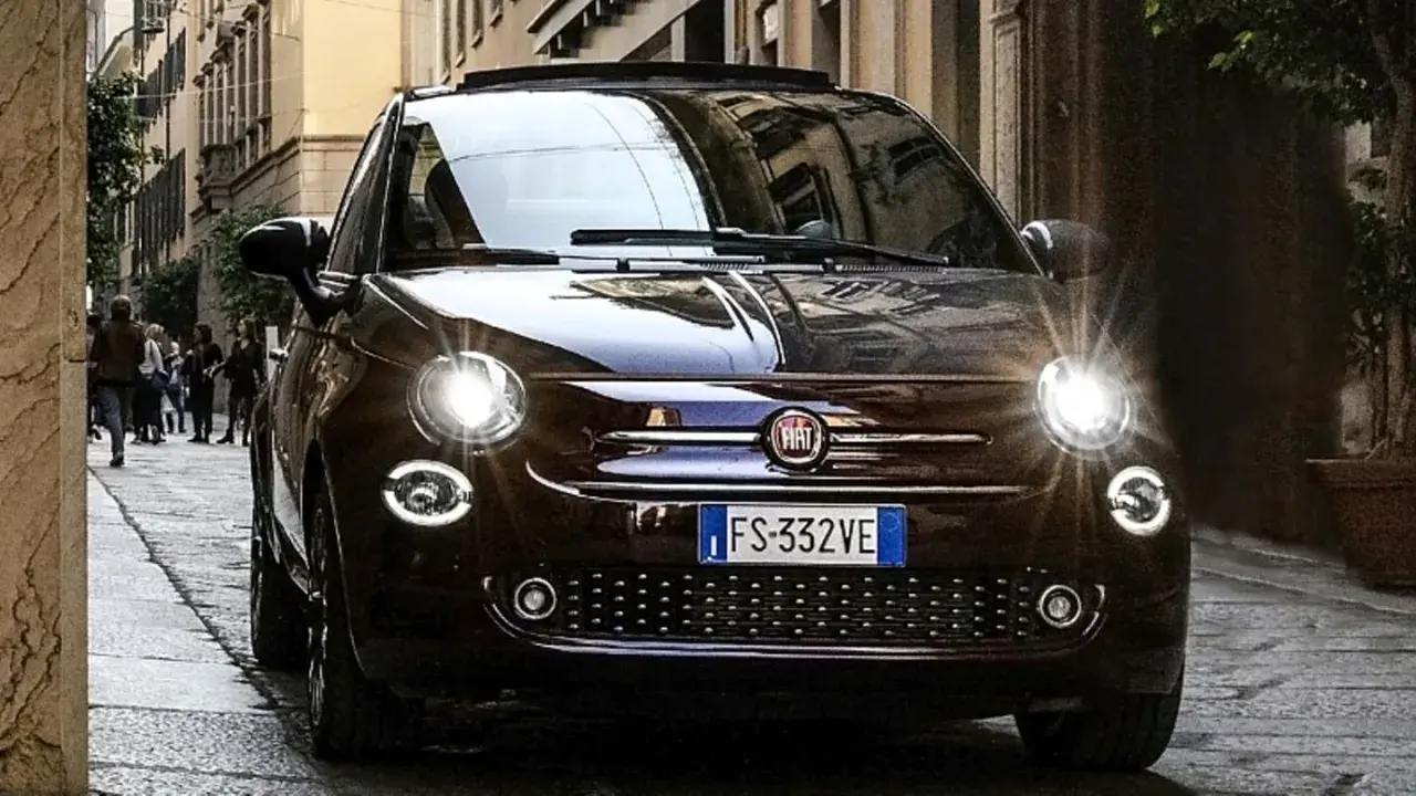 Dunkelbrauner Fiat 500 mit leuchtenden Scheinwerfern auf einer Kopfsteinpflasterstraße. Ein paar Leute gehen im Hintergrund vorbei. Keine sichtbaren Fiat 500 Probleme.