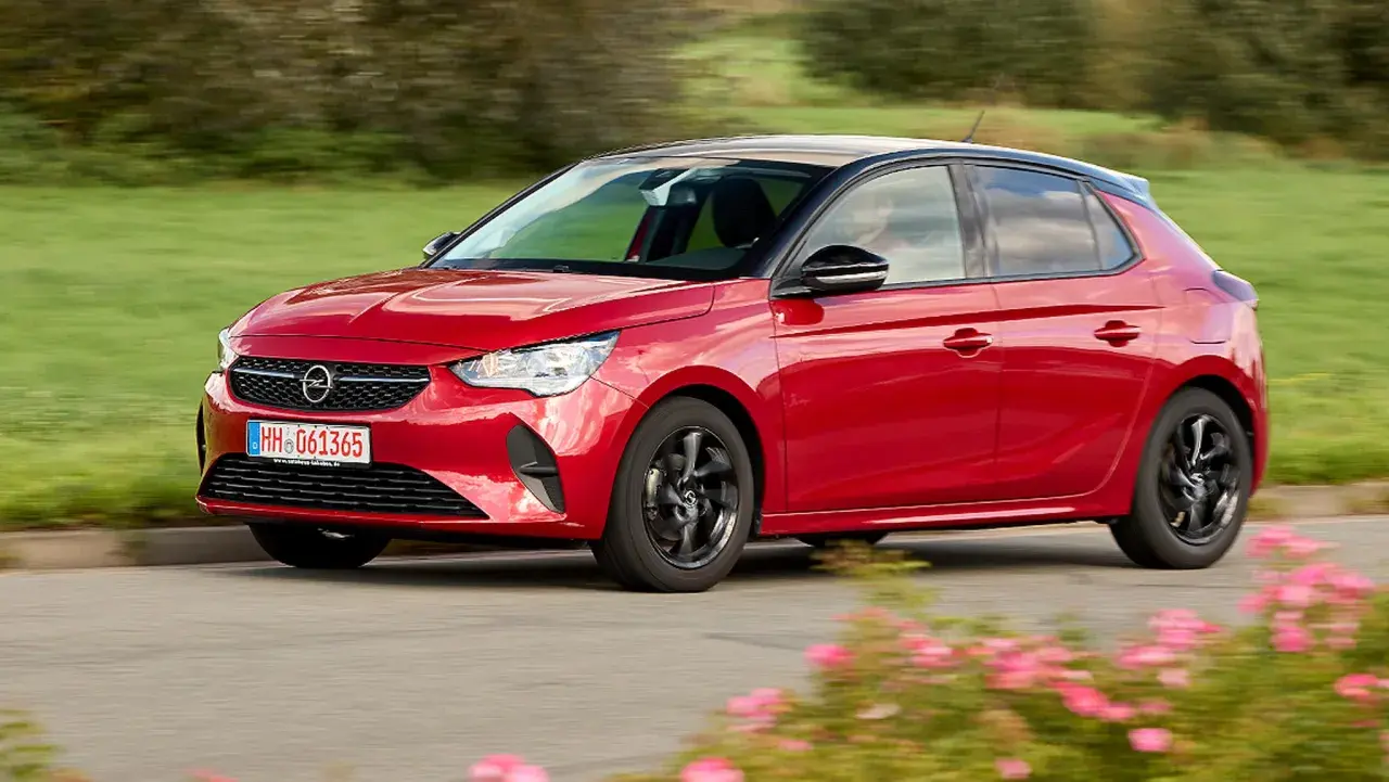 Roter Opel Corsa F fährt auf einer Straße, umgeben von grünen Feldern und blühenden Blumen. Keine Probleme mit diesem flotten Flitzer!