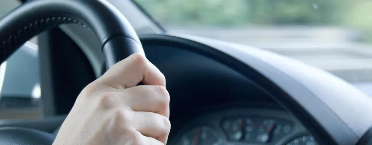 Hand am Lenkrad, das leicht flattert. Die Straße zieht vorbei, ein Gefühl von Bewegung.