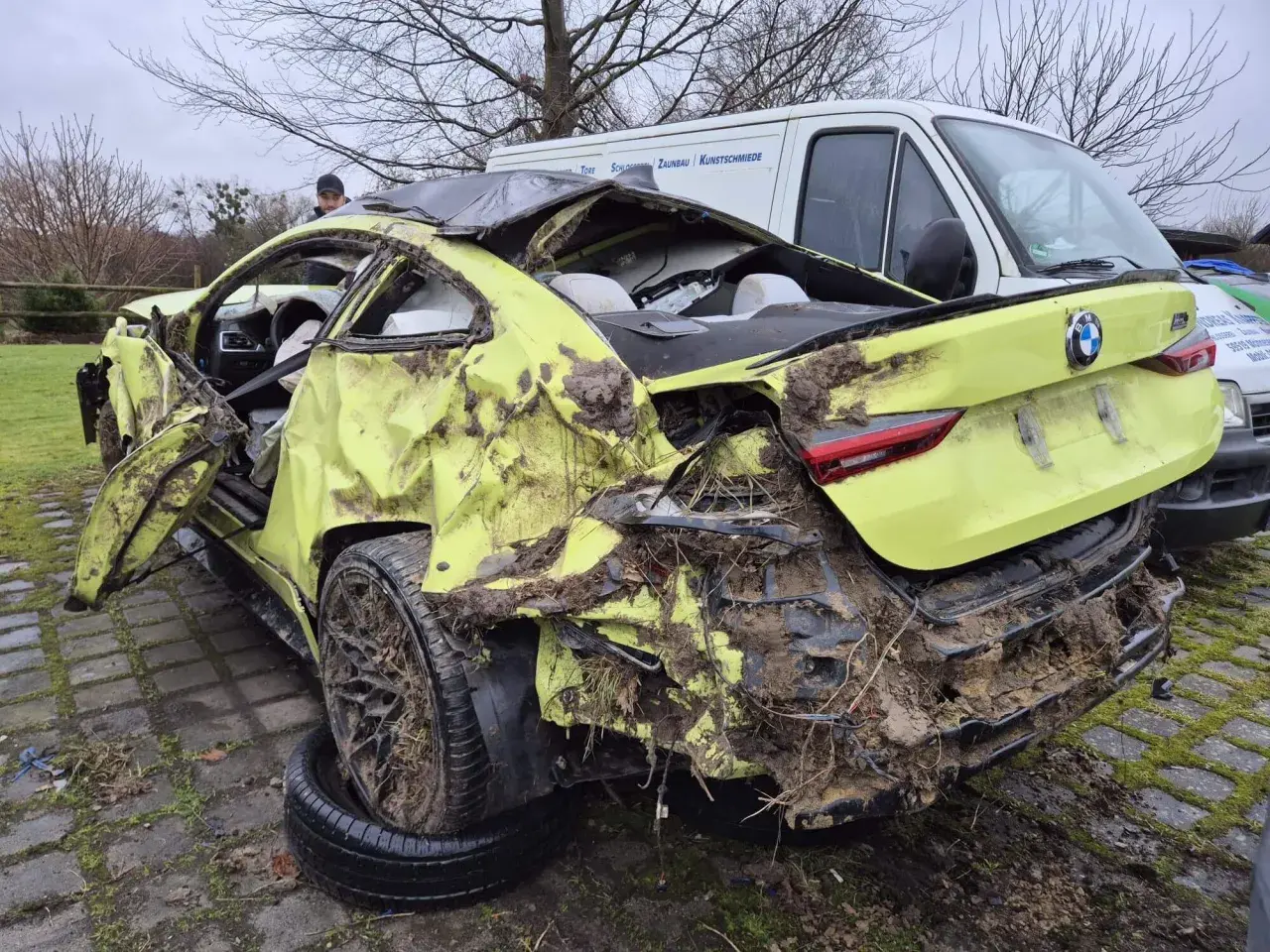 Ein gelber BMW mit Totalschaden, stark beschädigt und mit Schlamm bedeckt, steht neben einem weißen Lieferwagen.