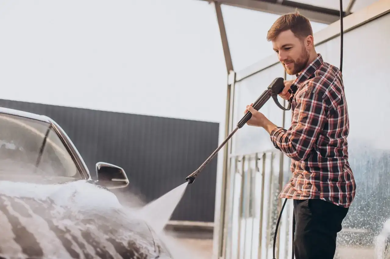 Ein Mann mit kariertem Hemd nutzt einen Hochdruckreiniger, um sein Auto zu waschen.