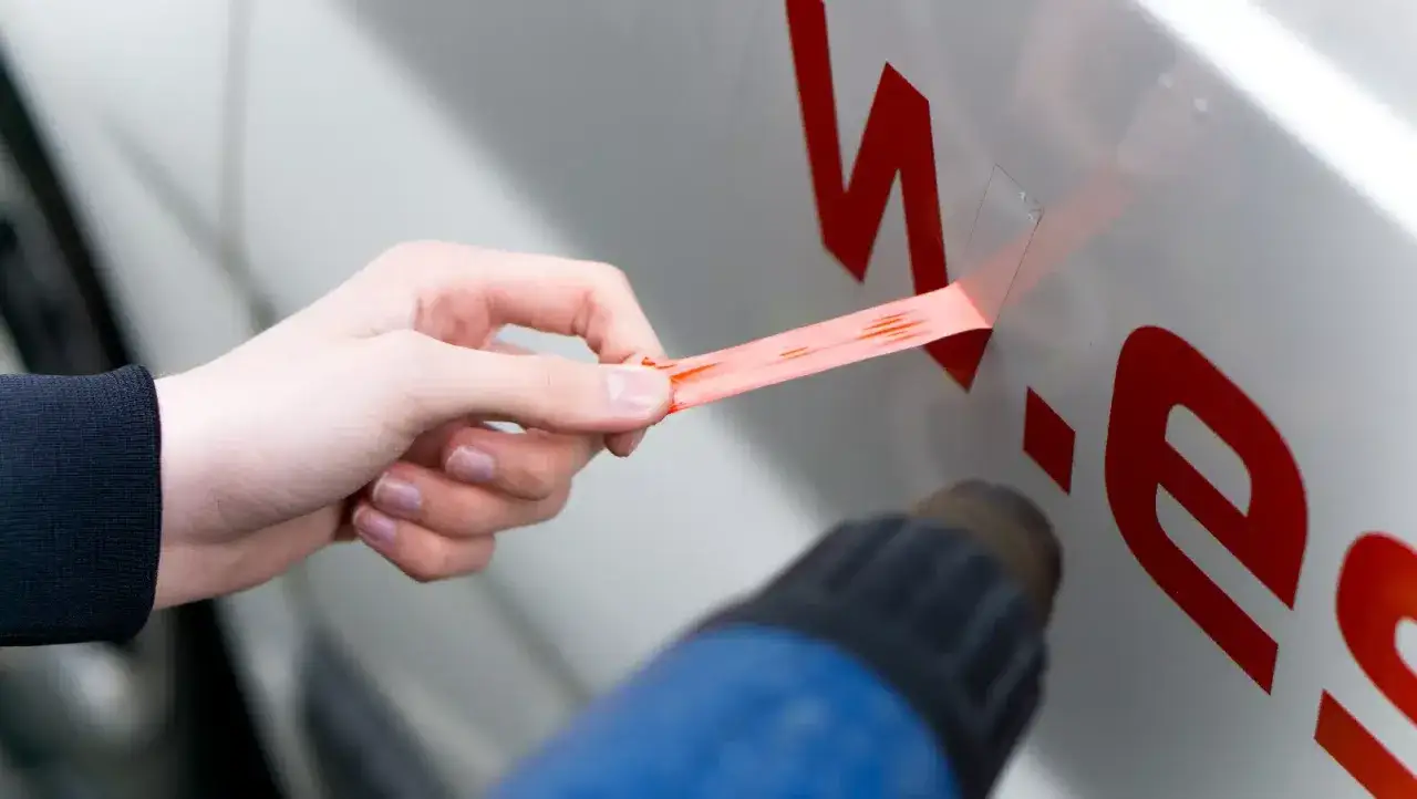 Hand zieht roten Aufkleber vom Auto ab, w&auml;hrend Hei&szlig;luftf&ouml;hn das Material erw&auml;rmt.