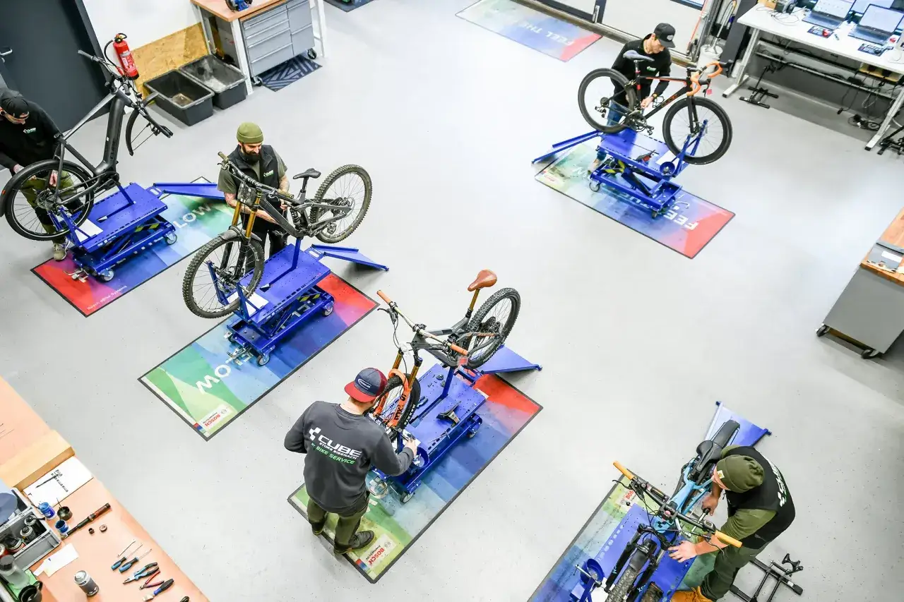 Mehrere Mechaniker f&uuml;hren Fahrrad Wartung in einer Werkstatt durch. Sie arbeiten an verschiedenen Mountainbikes und E-Bikes auf blauen Montagest&auml;ndern.