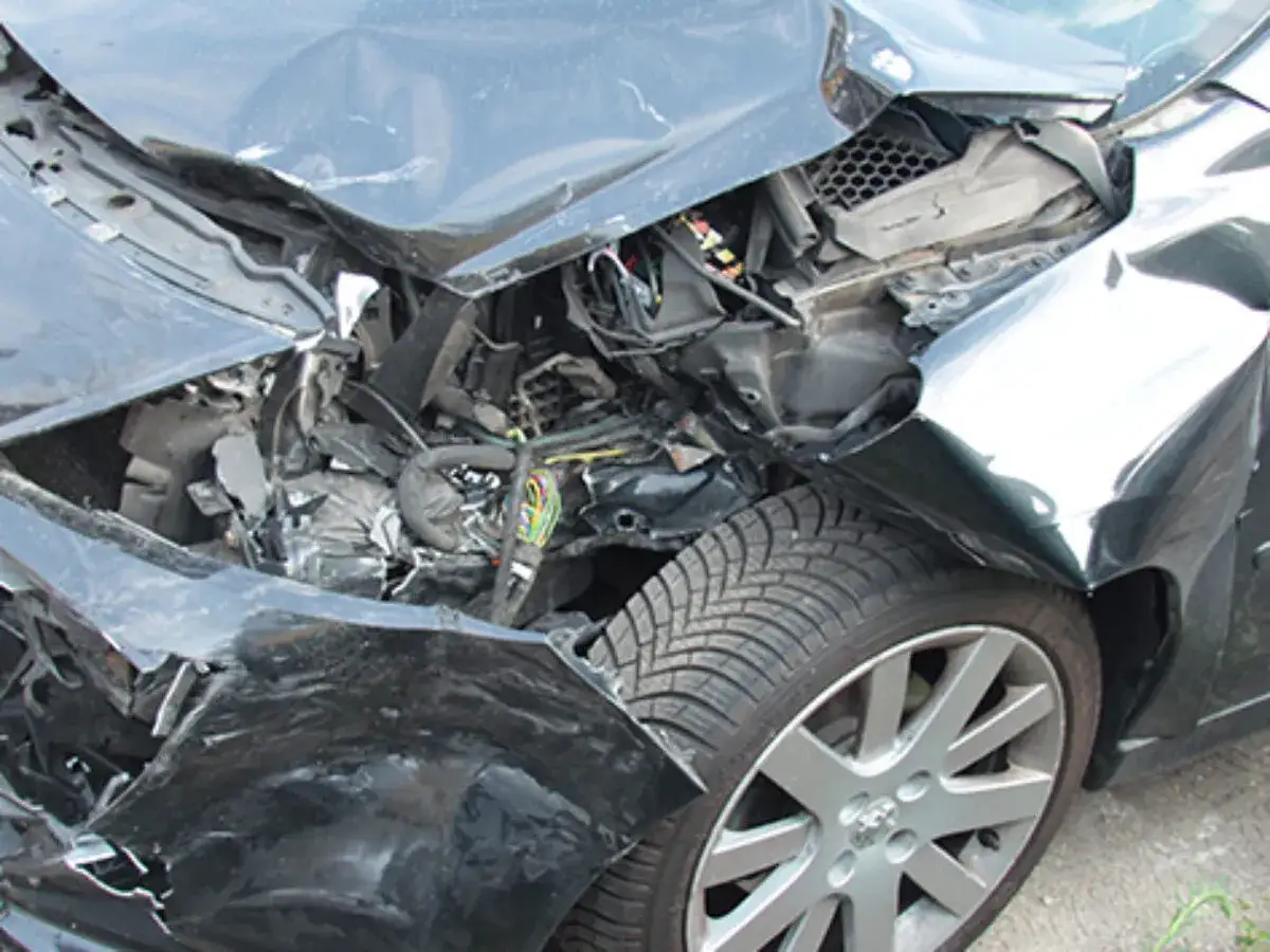 Ein schwarzes Auto mit Totalschaden nach einem Unfall. Die Front ist stark verformt, der Motorraum liegt frei, Kabel und Teile sind zerstreut.