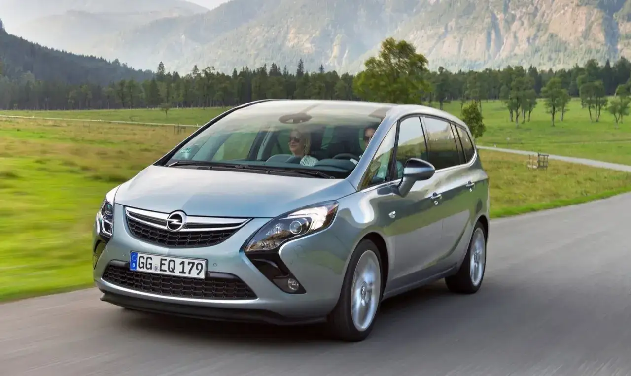 Silberner Opel Zafira C f&auml;hrt auf einer Landstra&szlig;e durch eine gr&uuml;ne Landschaft mit Bergen im Hintergrund.