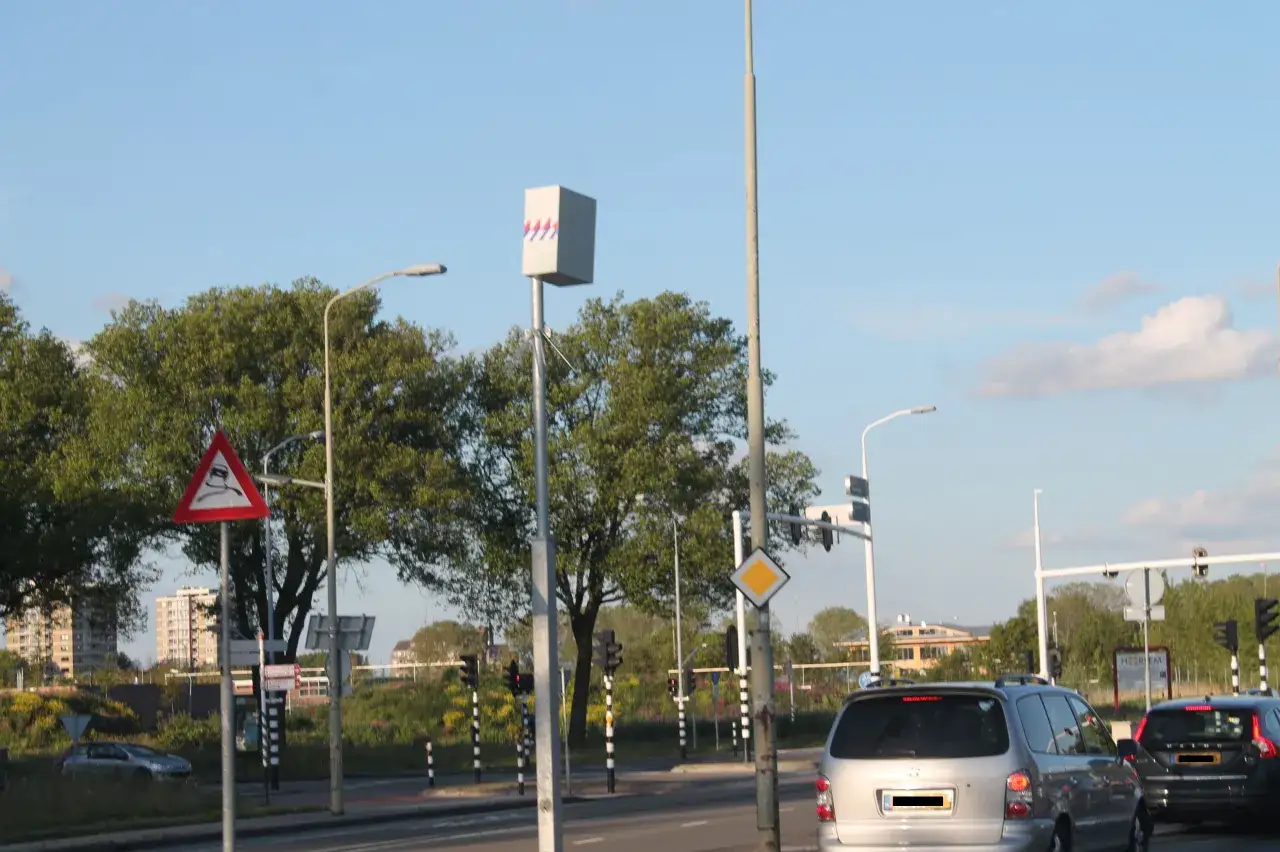 Ein Blitzer in den Niederlanden, auf einem Mast montiert, &uuml;berwacht den Verkehr. Verkehrsschilder warnen vor rutschiger Fahrbahn.