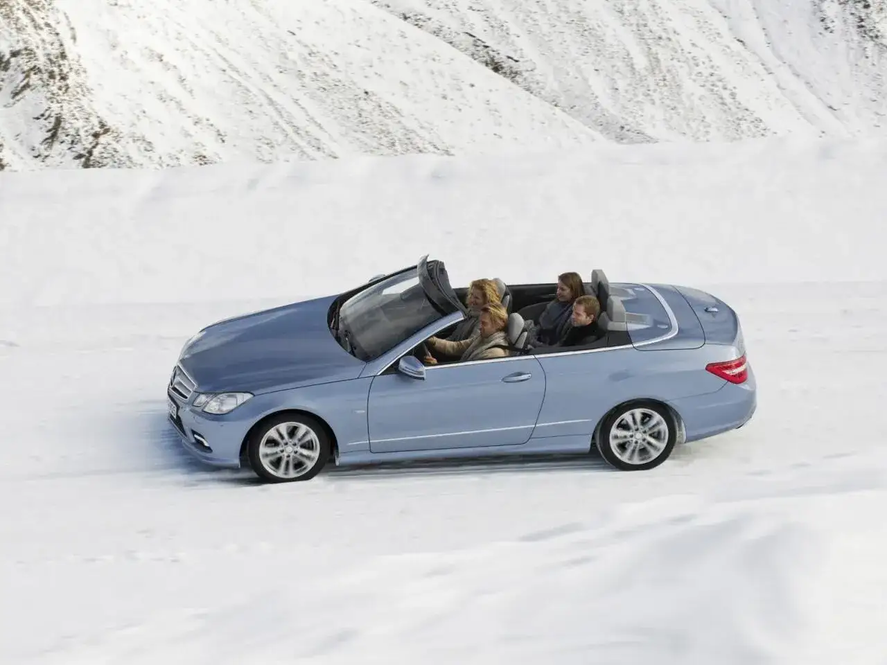 Ein blaues Cabrio f&auml;hrt durch eine verschneite Landschaft. Vier Personen genie&szlig;en die Fahrt im Freien, trotz des Winters.