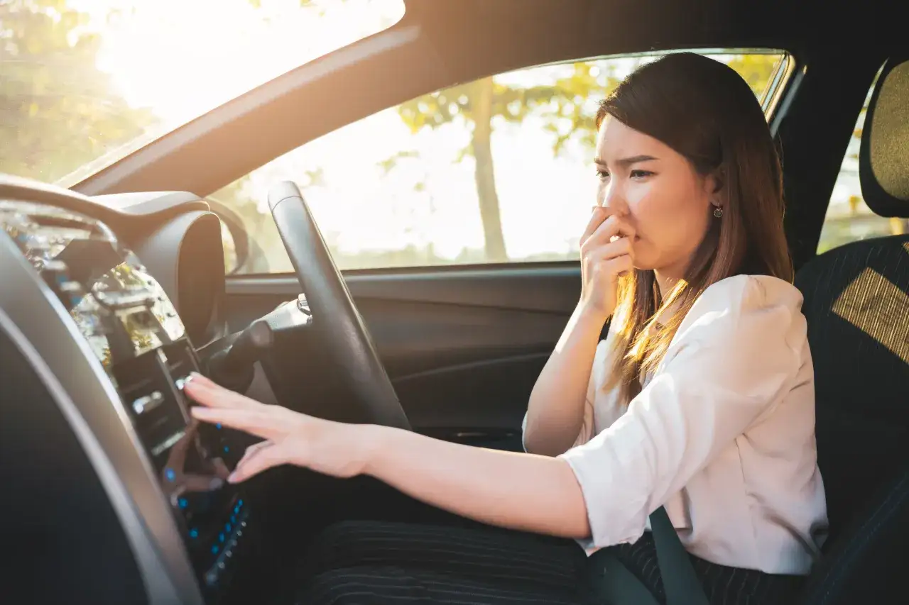 Frau im Auto h&auml;lt sich die Nase zu, weil das Auto nach Benzin riecht. Sie versucht, die L&uuml;ftung einzustellen.