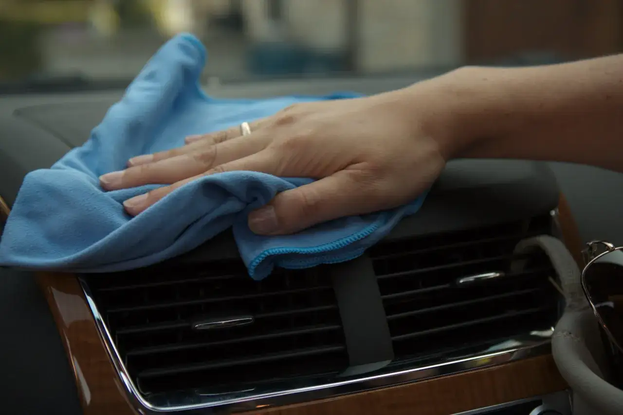 Hand wischt mit blauem Tuch &uuml;ber L&uuml;ftungsschlitze im Auto, um Staub zu entfernen.