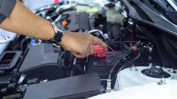 Hand mit Uhr lädt die Autobatterie ohne Abklemmen. Roter Stecker wird auf den Pluspol gesetzt.