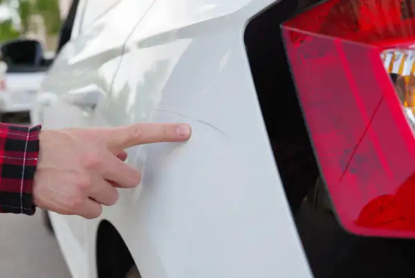 Finger zeigt auf Kratzer am weißen Auto, vermutlich verursacht durch die Waschanlage.