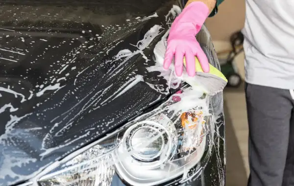 Eine Hand in pinker Handschuh-Hülle reinigt mit einem Schwamm das nasse, schwarze Auto. Schaum bedeckt die Motorhaube und den Scheinwerfer.