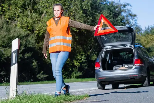 Frau mit Warnweste und Warndreieck neben offenem Kofferraum. Sie bereitet sich auf das auto aufbocken vor, um das Fahrzeug zu reparieren.