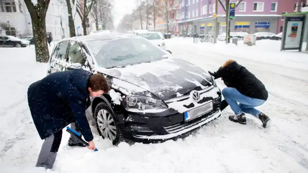 Zwei Personen schaufeln Schnee vom auto im winter. Ein Mann mit blauer Bürste reinigt das Rad, eine Frau schiebt die Motorhaube.