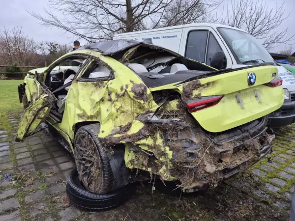 Ein gelber BMW mit Totalschaden, stark beschädigt und mit Schlamm bedeckt, steht neben einem weißen Lieferwagen.